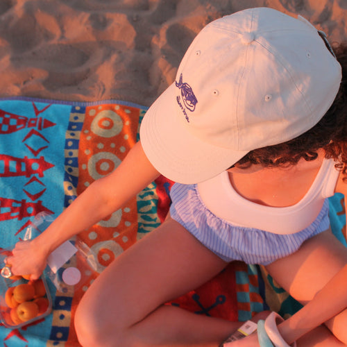 Woman sitting on a colourful towel at the beach wearing a beige embroidered cap. The perfect summer evening. 